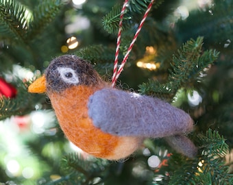May include: A felt robin ornament with a gray back, orange breast, and black head. The ornament is hanging from a red and white string on a green Christmas tree.