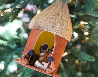 May include: A brown and tan thatched roof hut ornament with a yellow interior and three figures inside. The figures are black and are dressed in brown and white clothing. The ornament is hanging from a Christmas tree.