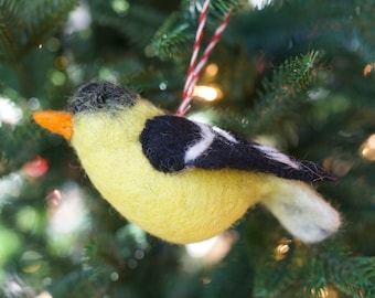 May include: A yellow and black felt bird ornament with a white stripe on its wing. The ornament is hanging from a red and white string on a green Christmas tree.