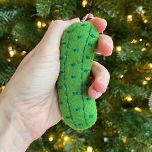 May include: A green felt pickle Christmas ornament with dark green embroidered details. The ornament has a red and white string for hanging. The background is a blurred Christmas tree.