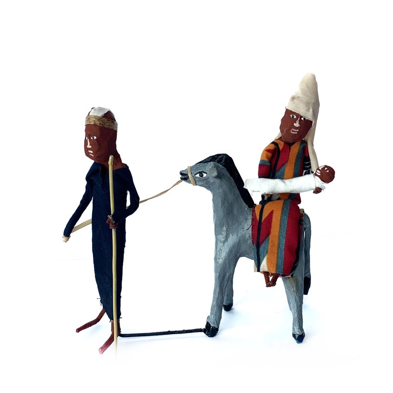 May include: Two hand-crafted figurines depicting a man on a donkey and a man walking with a staff. The figures are made of fabric and painted in brown, gray, white, red, and orange.