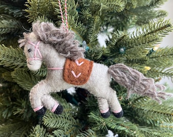 Może przedstawiać: Szary filcowy ornament w kształcie konia z brązowym filcowym siodłem i różowymi filcowymi akcentami. Koń jest zawieszony na czerwono-białej sznurce na choince.