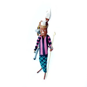 May include: A handmade doll with a pink and black striped shirt, blue and green patterned pants, and a white headdress. The doll is holding a wooden stick and has a brown face with painted features.