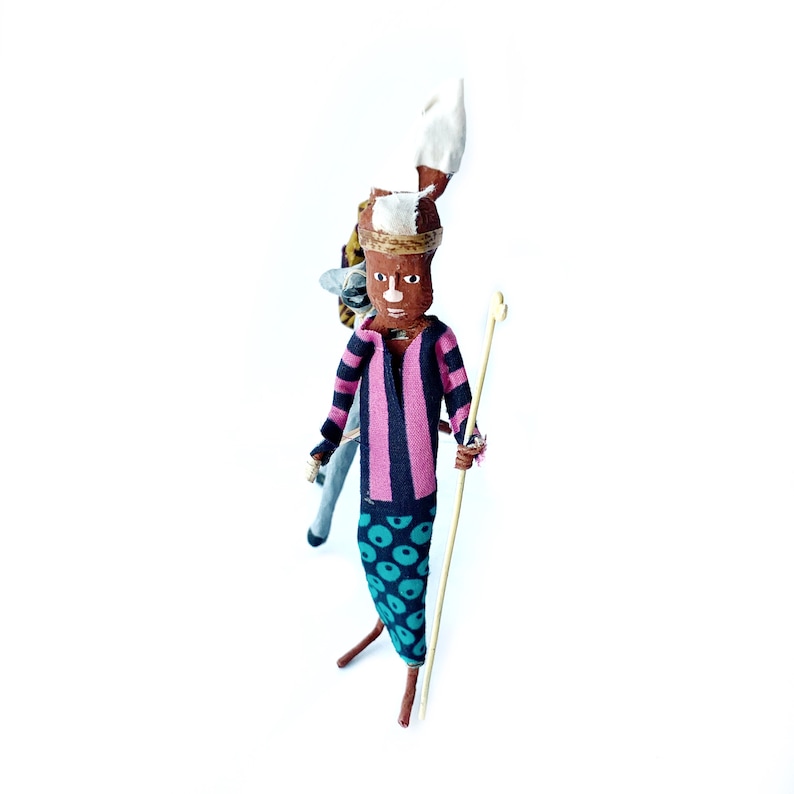 May include: A handmade doll with a pink and black striped shirt, blue and green patterned pants, and a white headdress. The doll is holding a wooden stick and has a brown face with painted features.