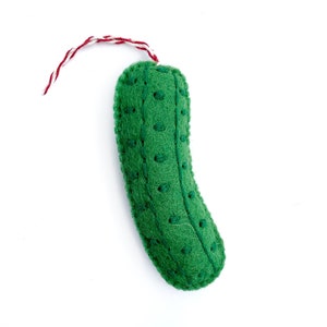 May include: A green felt pickle-shaped ornament with embroidered details and a red and white hanging string. The pickle has a curved shape and small, evenly spaced embroidered dots. The ornament is set against a white background.
