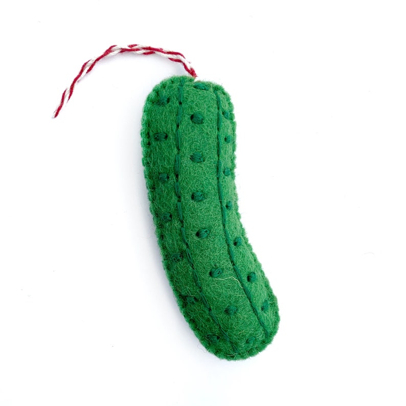 May include: A handmade, green felt pickle ornament with embroidered details and a red and white string for hanging. The pickle shape is curved, with small, evenly spaced embroidered dots. The ornament is set against a white background.