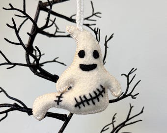 May include: A handmade white felt ghost ornament with black stitched details, including eyes, a mouth, and a stitched body. The ghost is hanging from a white string against a backdrop of black branches, perfect for Halloween decor.