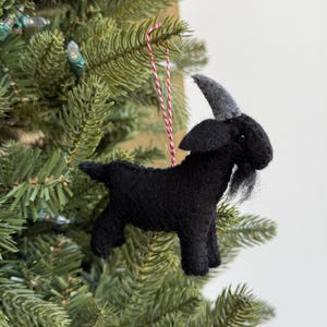 May include: A black felt goat Christmas ornament with grey horns and a long beard, suspended by a red and white string. The goat is set against a green Christmas tree, creating a festive holiday scene.