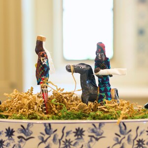 May include: Two handmade figurines, one riding a gray horse, are displayed in a white ceramic bowl with a blue and white floral pattern. The figurine on the horse is wearing a blue and green patterned robe and a red hat. The other figurine is wearing a blue and red patterned robe and a white hat. The bowl is filled with brown shredded paper and green moss.