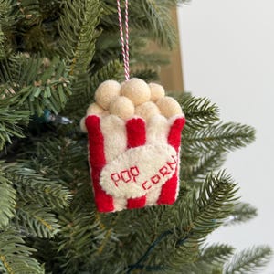 May include: A felt popcorn Christmas tree ornament. The popcorn container has red and white stripes, with the words "POP CORN" embroidered on the front. The popcorn is made of small, round, beige felt balls. It hangs from a red and white string.