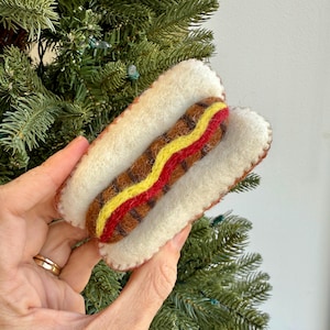May include: A felted hot dog ornament with a brown sausage, yellow mustard, and red relish, nestled in a white bun. The ornament is hand-stitched with brown thread and is a festive holiday decoration.