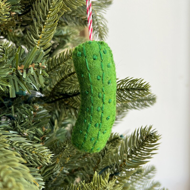 May include: A green felt pickle Christmas ornament with embroidered details and a red and white string for hanging. The ornament is hanging on a green Christmas tree.