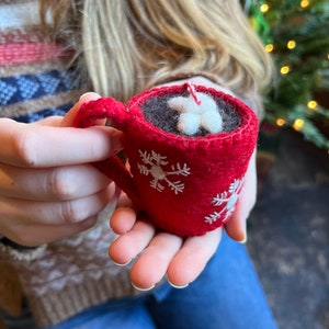 Girl holding hot chocolate ornament in front of Christmas tree.