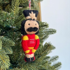 May include: A felt Christmas ornament shaped like a nutcracker. The nutcracker is wearing a red and yellow uniform with a black hat and mustache.