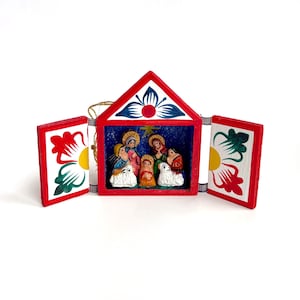 Puede incluir: Un pequeño adorno de madera de la natividad con un diseño pintado en rojo, blanco y verde. El adorno tiene una puerta con bisagras que se abre para revelar una escena de la natividad en miniatura con figuras de María, José, el niño Jesús y una oveja.