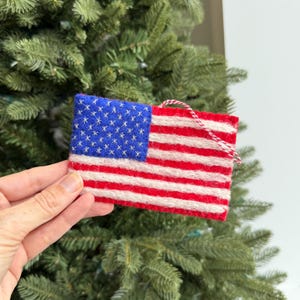 Puede incluir: Adorno rectangular de fieltro que representa la bandera estadounidense. Presenta un cuadrado azul con estrellas blancas en la esquina superior izquierda y rayas rojas y blancas. Se adjunta una cuerda roja y blanca para colgarlo. Adorno navideño.