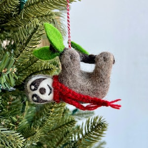 May include: A brown and gray felt sloth ornament hanging from a red and white string. The sloth is wearing a red knitted scarf and is holding onto a green felt branch.