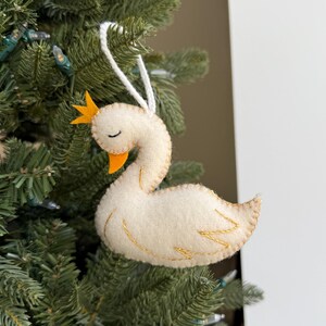 May include: A handcrafted Christmas tree ornament shaped like a swan. The swan is made of off-white felt with gold stitching, an orange beak, and a yellow crown. It hangs from a white string against a backdrop of green Christmas tree branches.