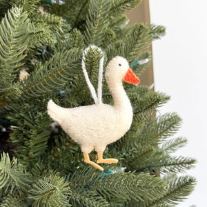 May include: A cream-colored goose ornament with an orange beak and yellow feet hangs on a green Christmas tree. The goose is made of a soft, textured material and has a white string for hanging. A festive holiday decoration.