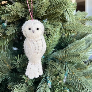 May include: A white owl ornament with black eyes and a red and white string for hanging. The owl is made of a soft, textured material and is decorated with small, dark stitches. The ornament is hanging on a green Christmas tree.