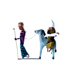 May include: A handmade nativity scene featuring three figures. A man with a staff walks beside a donkey carrying a woman holding a baby. The figures are made of fabric and have painted faces.