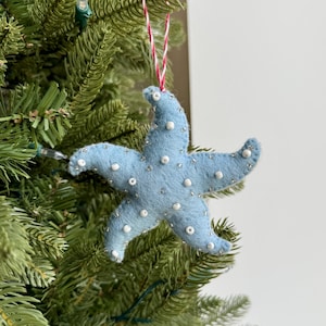 May include: A light blue felt starfish Christmas ornament with white beaded accents. The ornament is hanging from a red and white string and is displayed on a green Christmas tree. The starfish has five points and is decorated with small, round white beads.