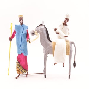 May include: Two figures made of paper mache, one riding a donkey. The figure on the donkey is wearing a white robe and a white turban. The other figure is wearing a blue robe with a red and gold patterned skirt and a gold crown. The donkey is gray with a brown saddle and a yellow rope bridle.