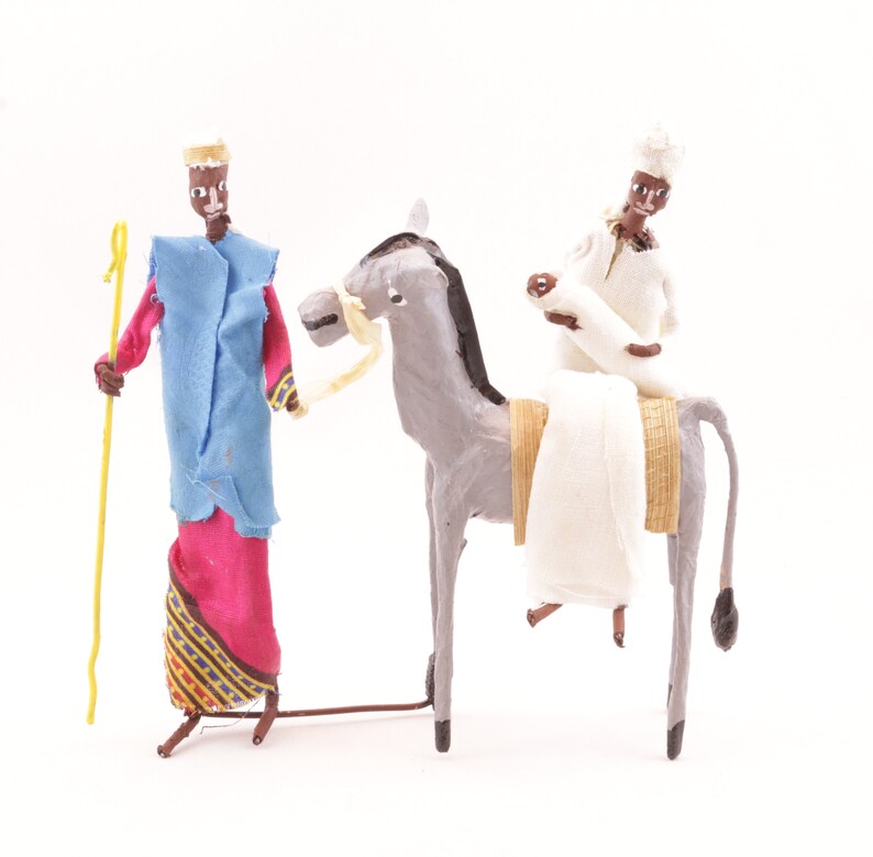 May include: Two figures made of paper mache, one riding a donkey. The figure on the donkey is wearing a white robe and a white turban. The other figure is wearing a blue robe with a red and gold patterned skirt and a gold crown. The donkey is gray with a brown saddle and a yellow rope bridle.