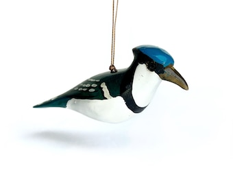 May include: A hand-painted wooden bird ornament. The bird features a blue head, a white chest, and a dark green back with white spots. It has a brown beak and is suspended by a thin, brown string. The ornament is designed to resemble a blue jay.