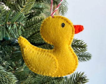Puede incluir: Un adorno de pato de fieltro amarillo con un pico rojo y ojos negros. El adorno está colgado de una cuerda roja y blanca en un árbol de Navidad verde.