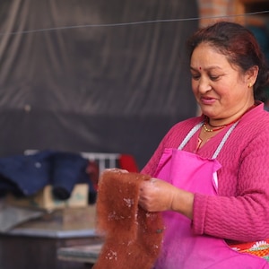 May include: A woman wearing a pink apron and a pink sweater holds a brown piece of fabric in her hands. She is working in a workshop with a variety of colorful fabrics in the background.
