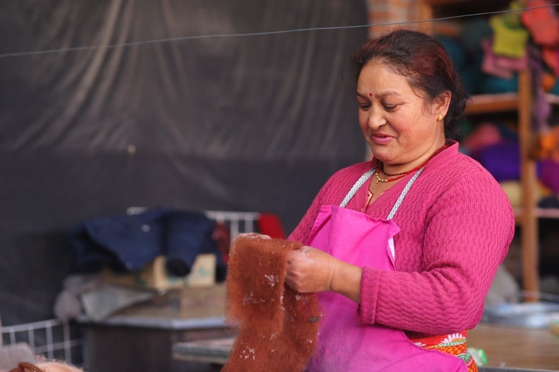 May include: A woman wearing a pink apron and a pink sweater holds a brown piece of fabric in her hands. She is working in a workshop with a variety of colorful fabrics in the background.