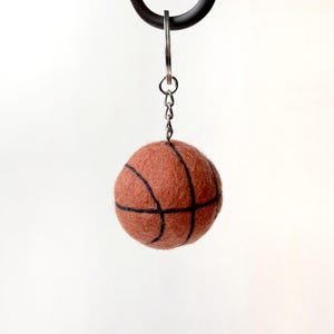 May include: A brown basketball keyring with black lines, suspended from a silver chain and ring. The felt ball is round and resembles a miniature basketball. A sports-themed accessory.