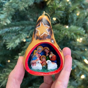 May include: A hand-painted Christmas ornament shaped like a gourd, featuring a nativity scene inside. The ornament has a star design and a red border. The background is a Christmas tree.