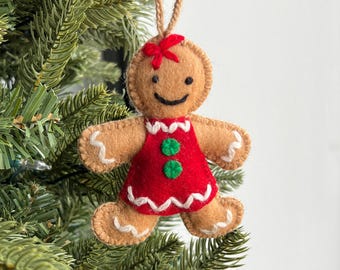 May include: Felt gingerbread person ornament with a red dress, white trim, and green buttons. The ornament has a brown face with a smile and a red bow on top of its head.