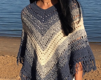 Puede incluir: Un poncho de crochet azul y blanco con flecos. El poncho lo lleva puesto una persona con el pelo largo y negro. La persona está de pie en una playa de arena con una masa de agua al fondo.