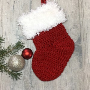Peut inclure: Chaussette de Noël rouge en crochet avec un revers en fausse fourrure blanche. La chaussette est sur un fond en bois avec deux boules argentées et rouges.