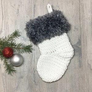Peut inclure: Chaussette de Noël en crochet blanc avec un revers en fausse fourrure grise. La chaussette est accrochée sur un fond en bois avec une branche de pin et des décorations.