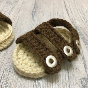 May include: A pair of brown and beige crocheted baby sandals with silver buttons.