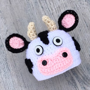 May include: Crochet cow hat pattern. The hat is white with black spots and pink ears. It has googly eyes and a black button nose.