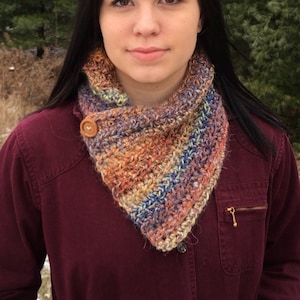 Puede incluir: Una mujer con una camisa granate con una bufanda de cuello de chal de ganchillo en tonos naranja, marrón, azul y blanco. La bufanda tiene un botón de madera.