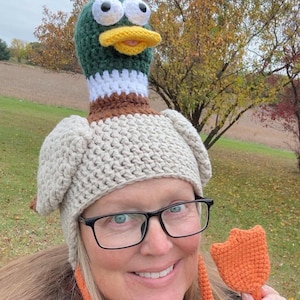 Könnte beinhalten: Handgefertigte Häkel-Entenmütze mit grünem Kopf, gelbem Schnabel und weißen Augen. Die Mütze hat einen beigen Körper und Ohrenklappen. Eine orangefarbene gehäkelte Entenfuß wird hochgehalten. Ideal für Kostümpartys oder lustige Veranstaltungen.