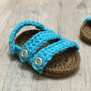 May include: A pair of crocheted baby sandals with blue straps and brown soles. The sandals have silver buttons.