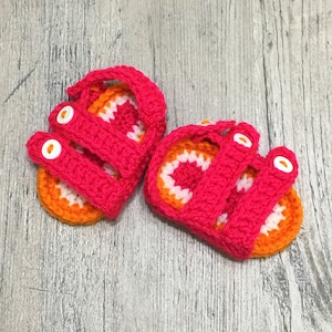 May include: A pair of crocheted baby sandals in pink and orange with white buttons. The sandals have a strap design and are made with a soft, textured yarn.