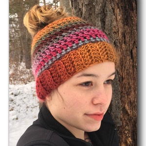 May include: A woman wearing a colorful crocheted hat with a bun on top. The hat has stripes of orange, brown, pink, gray, and red.