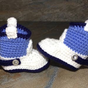 May include: A pair of handmade crochet baby cowboy boots. The boots are white with blue accents and a dark blue trim. Each boot has a silver-colored spur and a button detail. The boots are designed to look like miniature cowboy boots.