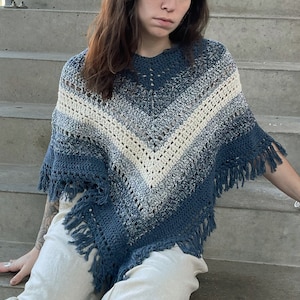 May include: A blue and white crocheted poncho with fringe. The poncho is being worn by a person sitting on a set of concrete steps.