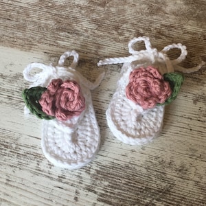May include: A pair of white crocheted baby sandals with pink and green flower embellishments.