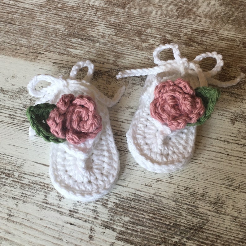 May include: A pair of white crocheted baby sandals with pink and green flower embellishments.
