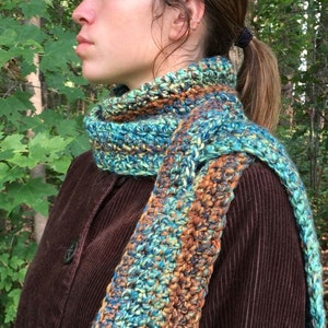 May include: A crocheted scarf with a blue, green, and brown color pattern. The scarf is worn around the neck of a person wearing a brown corduroy jacket.